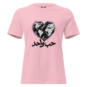 May include: A light pink short-sleeve t-shirt featuring a graphic of a heart-shaped world map in black and white. Below the heart, the Arabic text "حب واحد" is printed in bold black calligraphy, centered on the chest of the garment.