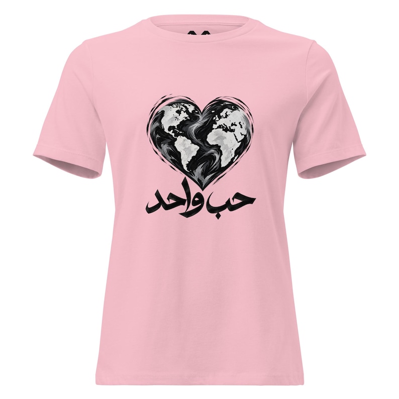 May include: A light pink short-sleeve t-shirt featuring a graphic of a heart-shaped world map in black and white. Below the heart, the Arabic text "حب واحد" is printed in bold black calligraphy, centered on the chest of the garment.