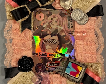 Könnte beinhalten: Ein rechteckiger Kartenrahmen, verziert mit rosa Spitze, goldfarbenem Glitzerband sowie schwarzen und rosa Satinschleifen. Der Rahmen enthält eine holografische Anime-Charakterkarte, einen rosa Herzanhänger, eine schwarze Rose und einen Filmstreifen.