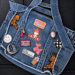 May include: A blue denim tote bag decorated with various embroidered patches including two tigers, a frog, a pink balloon dog, a cassette tape, a coffee cup, a smiley face, and a rectangular patch with the text It's fine. I'm fine. Everything's fine.