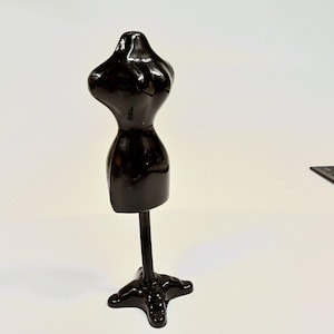 May include: A glossy black miniature dress form mannequin featuring a smooth, contoured torso shape mounted on a slender central pole and a stable, three-pronged base. The object has a reflective surface and a solid, uniform dark color throughout.