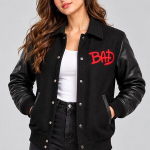 May include: A black varsity jacket featuring long black sleeves, a collared neckline, and a front button closure. The word BAD is embroidered in red thread on the upper left chest area. The jacket includes ribbed cuffs and a ribbed hemline.
