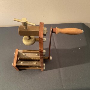 May include: A manual pasta machine featuring a wooden frame, brass components, and a metal crank handle with a wooden grip. The device includes a top-mounted screw mechanism with a T-shaped handle and a lower section with two parallel metal rollers.