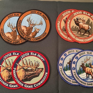 May include: A collection of eight circular embroidered patches featuring elk imagery and text. The patches are dated 2001, 2002, 2003, and 2005, each labeled Pennsylvania Game Commission and Elk Hunt, with varying border colors including black, red, and blue.