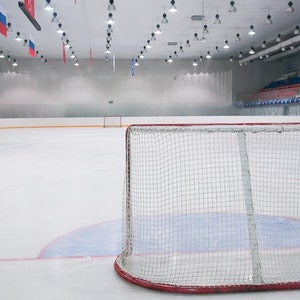May include: An empty hockey rink with a white surface and red and yellow lines. A red and white hockey net is in the foreground.