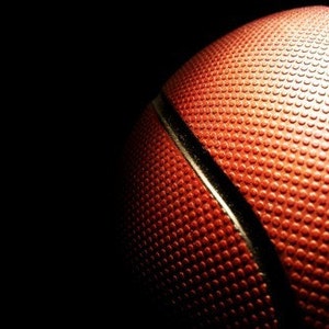May include: Close-up of a basketball, showcasing its textured orange surface and black seams. The ball is partially lit against a dark background, highlighting the dimpled texture and the classic design of the sports equipment.