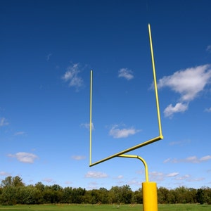 May include: A yellow metal football goal post against a bright blue sky with white clouds.