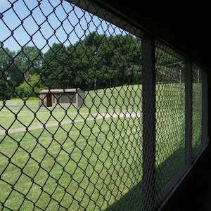 May include: View of a baseball field through a chain-link fence. The field is green with a small dugout and trees in the background. The sky is blue and the day is sunny.