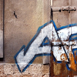 May include: A close-up of a rusty metal door with white and blue graffiti. The door is partially open and has a metal latch. The wall behind the door is a light brown color.