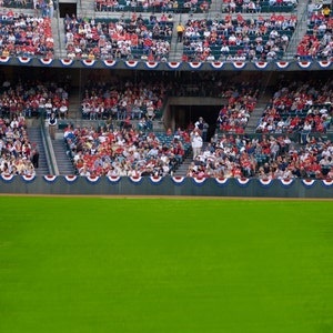 May include: A baseball stadium filled with spectators. The stands are packed with people wearing red and white. An American flag waves in the sky above the stadium. Bright stadium lights are visible.