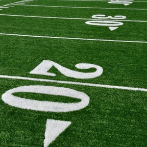 May include: A green artificial turf football field with white yard lines and numbers. The numbers 20 are visible in the foreground.