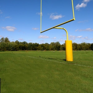 May include: A yellow metal football goal post stands on a grassy field against a blue sky with white clouds.