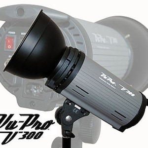May include: A gray and black Vu-Pro V-300 studio flash with a black reflector. The flash has a power switch and a mode dial. The flash is mounted on a black stand.