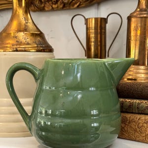 May include: A sage green ceramic pitcher with a rounded, ribbed body and a single curved handle. The pitcher features a smooth, glossy finish and a distinct pouring spout, showcasing a simple, functional design with horizontal ridges.