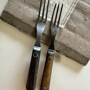 May include: Two antique forks with dark wooden handles and metal tines. One handle is dark brown, the other is light brown. The forks are resting on a textured, aged paper surface, suggesting a vintage or rustic aesthetic.