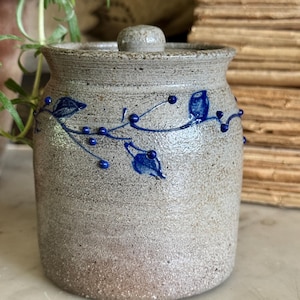 May include: A cylindrical stoneware jar with a speckled light gray finish and a matching lid. The exterior features a hand-painted cobalt blue vine motif with small leaves and berries encircling the upper portion of the vessel body.