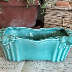 May include: A rectangular turquoise ceramic planter with a glossy finish and vertical fluted detailing along the sides. The vessel features a scalloped, wavy rim and a slightly curved base, showcasing a vibrant, solid color throughout the exterior.