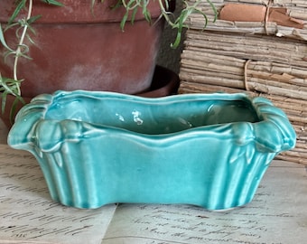 Puede incluir: Una maceta rectangular de cerámica turquesa con acabado brillante y detalles estriados verticales en los laterales. El recipiente presenta un borde festoneado y ondulado con una base ligeramente curva, mostrando un color sólido y vibrante.
