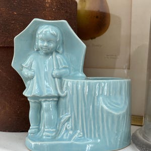 May include: A light blue ceramic planter featuring a young girl holding an umbrella. The planter has a tree trunk design with a draped fabric detail. A charming vintage piece for home decor.