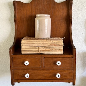 May include: A small, antique wooden wall shelf with a decorative back and two drawers. The shelf holds a stack of aged paper tied with twine and a beige ceramic jar. The drawers have white, round knobs.