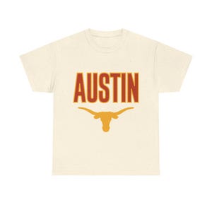 Könnte beinhalten: Ein cremefarbenes Kurzarm-T-Shirt mit gesprenkelter Textur. Auf der Vorderseite befindet sich das Wort AUSTIN in fetten roten Buchstaben mit einem dünnen gelben Umriss, platziert über einer gelben Grafik einer Longhorn-Stierkopf-Silhouette.