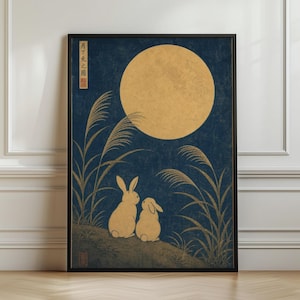 Op de afbeelding: Een ingelijste kunstdruk met twee lichtgekleurde konijnen die op een heuvel zitten onder een grote, gouden volle maan tegen een donkerblauwe achtergrond. Hoge, slanke grassen omlijsten het tafereel en verticale Japanse tekst is zichtbaar in de linkerbovenhoek.