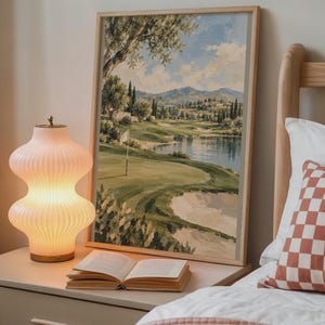 May include: A framed landscape painting depicting a golf course with a green fairway, a sand bunker, a small lake, and distant mountains under a blue sky with clouds. The artwork is rendered in a painterly style with visible brushstrokes.