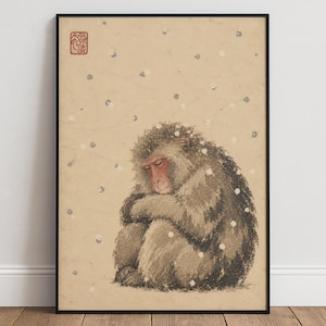Könnte beinhalten: Ein gerahmter Kunstdruck, der einen japanischen Makaken in zusammengerollter Haltung mit geschlossenen Augen zeigt. Der Affe hat dichtes, strukturiertes braunes Fell und ein rotes Gesicht. Kleine weiße Schneeflocken fallen vor beigem Hintergrund.