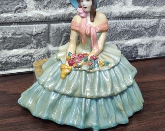 Peut inclure: Une figurine en porcelaine représentant une personne portant un chapeau bleu et une robe bleu clair à volants superposés. La figurine a une écharpe rose sur les épaules et tient un petit bouquet de fleurs multicolores sur ses genoux, finition brillante.