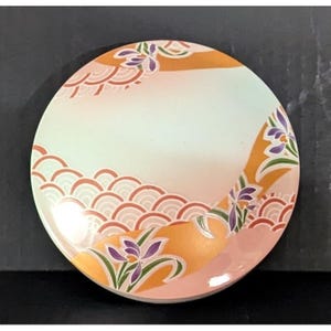 May include: A circular, flat decorative plate featuring a pale cream center with a gradient transition to soft pink. The design includes orange curved bands, stylized purple iris flowers with green leaves, and repeating orange wave patterns.