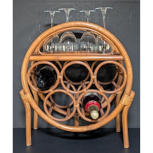May include: A circular light brown rattan wine rack featuring a top shelf for four stemmed glass goblets and a lower section with six circular openings for bottles. One dark glass bottle and one bottle with a red label are stored inside.