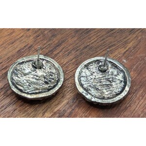May include: A pair of round stud earrings with a textured, antique silver-toned finish. Each earring features a circular metal base with a rough, uneven surface pattern and a central post backing, showcasing a weathered, metallic appearance.