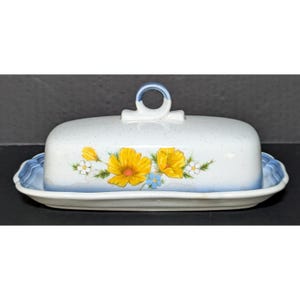 May include: A ceramic butter dish with a speckled white finish, featuring a central loop handle on the lid. The exterior is decorated with yellow flowers and small blue accents, while the base and lid edges are trimmed with a soft blue gradient.