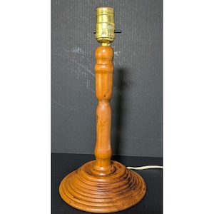 May include: A table lamp base crafted from light brown wood, featuring a smooth, turned central column and a wide, circular, tiered base. At the top, a metallic gold-colored light bulb socket is attached, complete with a small black switch.