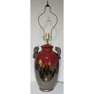 May include: A ceramic table lamp base featuring a glossy red glaze at the top that transitions into a dark, metallic brown drip pattern toward the bottom. The vessel has two curved handles on the sides and a brass-colored metal harp fixture.