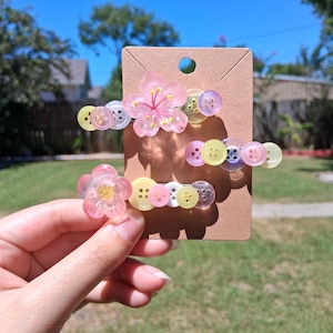 Puede incluir: Dos horquillas para el pelo sujetas a una tarjeta de cartón marrón. Cada horquilla presenta una flor rosa translúcida y una hilera de pequeños botones de plástico de colores en tonos amarillo, rosa, blanco y morado, dispuestos en línea.