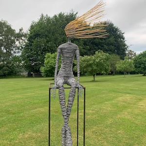 May include: A sculpture of a seated figure constructed from dark grey wire, featuring long limbs and a torso wrapped in tight coils. The head is composed of numerous thin, golden-yellow wire strands extending outward to one side like windblown hair.