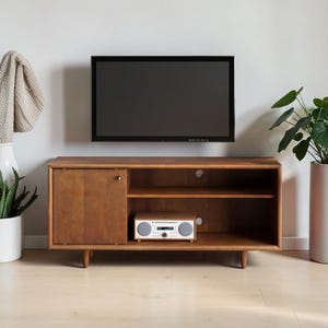 May include: A rectangular wooden media console with a single cabinet door on the left and an open shelf on the right. The console features four tapered legs and a light brown finish. A small silver-toned audio player sits on the shelf.