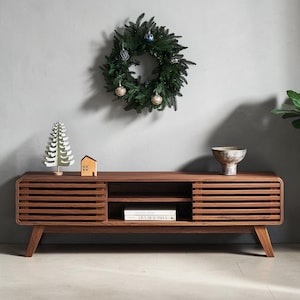 May include: A rectangular wooden media console featuring two slatted sliding doors on the sides and an open central shelving unit. The console has four tapered legs and holds a small paper tree, a wooden house, and a bowl on its top surface.