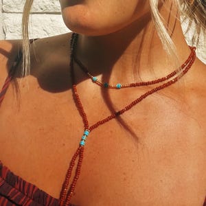 May include: A layered necklace featuring small rust-colored beads, accented with metallic gold beads and bright turquoise cylindrical spacers. The design includes a longer strand that hangs down in a lariat style, finished with turquoise bead accents.