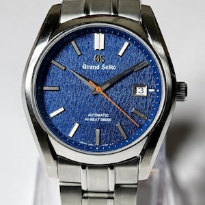May include: A stainless steel Grand Seiko watch with a textured blue dial, silver-tone hour markers, and a date window at the three o'clock position. The watch features a bright orange seconds hand, silver hour and minute hands, and a metal link bracelet.
