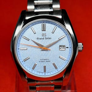 May include: A stainless steel Grand Seiko wristwatch with a textured light blue dial, silver-tone hour markers, and a date window at the three o'clock position. The watch features an orange second hand, silver hands, and a metal link bracelet.