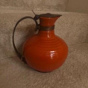 May include: A bright orange ceramic pitcher with a rounded body and horizontal ribbed texture. It features a dark metal handle attached to a metal band around the neck, topped with a matching metal lid that covers the spout opening.