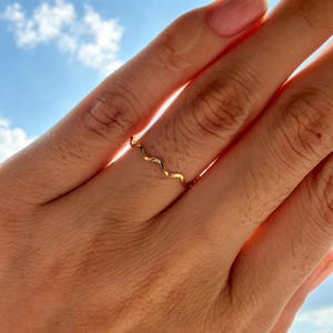 May include: A thin, gold-coloured metal ring featuring a continuous, delicate wavy or serpentine design. The band has a smooth, polished finish that reflects light, creating a simple and uniform aesthetic across the entire circumference of the ring.