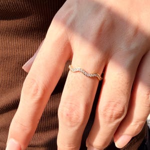 Può includere: Un anello color oro caratterizzato da un delicato design a fascia ondulata. La superficie dell'anello è incastonata con una fila continua di piccole pietre chiare a taglio rotondo che seguono la forma sinuosa della fascia metallica, creando un effetto scintillante.