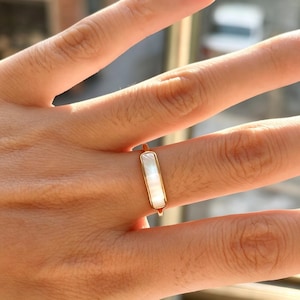 May include: A gold-toned ring featuring a slender, rectangular bar inlay made of iridescent white mother-of-pearl. The ring has a thin, polished metal band that encircles the finger, showcasing a clean, geometric design with a smooth finish.