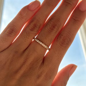 May include: A thin, metallic band ring featuring a continuous row of small, clear, round-cut stones set along the entire outer circumference. The ring has a polished finish and reflects light, showcasing a uniform design with multiple sparkling accents.