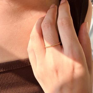 May include: A thin gold ring featuring a continuous band of small, uniform metal beads. The ring is worn on the middle finger, showcasing a delicate, textured design with a polished metallic finish that reflects light across its circular surface.