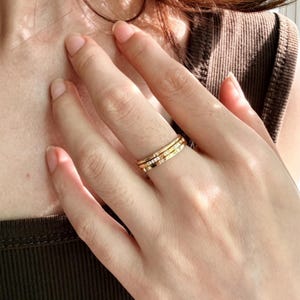 Può includere: Un set di tre anelli dorati sovrapposti indossati su un dito. L'anello superiore è una fascia sottile e liscia, quello centrale presenta una fila di piccole pietre chiare incastonate e quello inferiore è una fascia dorata più larga e lucida.
