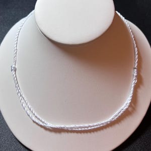 May include: A simple white cord necklace featuring a thin, twisted rope design. The necklace is constructed from a smooth, shiny white material and includes small, integrated sliding knots on each side to allow for adjustable length and fit.